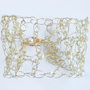 May include: A gold-coloured wire cuff bracelet with a delicate, open weave design. The bracelet is adorned with small, white pearls and features a gold clasp. The jewellery piece is set against a white background.