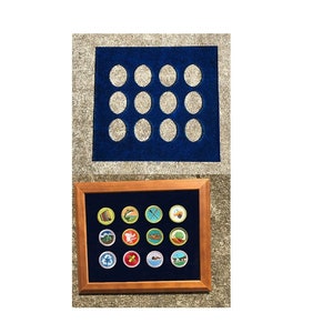 May include: A wooden frame with a blue velvet backing holds 16 colorful embroidered patches. The patches are arranged in a grid pattern and feature various designs, including a canoe, a cross, a tree, and a house. The frame is sitting on a gray concrete surface.
