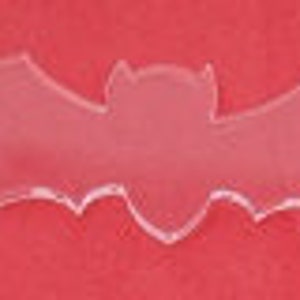 May include: A clear, bat-shaped ornament with outstretched wings. The bat is made of a transparent material and is set against a red background. The design is simple and symmetrical, with a pointed head and wing tips.