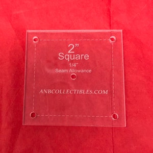 May include: A clear plastic 2-inch square quilting ruler with 1/4-inch seam allowance markings. The ruler has holes in each corner and is labeled "2" Square 1/4" Seam Allowance" and "ANBCOLLECTIBLES.COM".