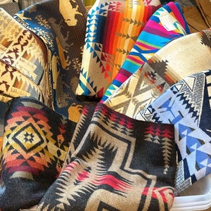 May include: A collection of colorful woven blankets with geometric patterns in shades of blue, red, yellow, brown, and white. The blankets are folded and stacked on top of each other.