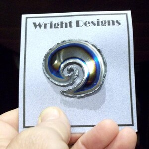 May include: A silver swirl design pin with blue and purple iridescent coloring. The pin is on a white card with the text "Wright Designs" printed in black.