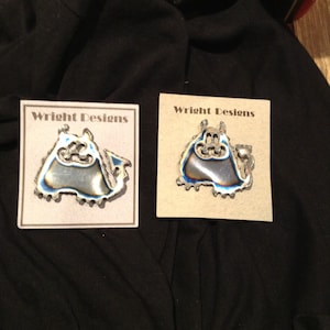 May include: Two silver metal pins with a blue and purple iridescent finish, shaped like a cartoon monster. The pins are on a beige card with the text "Wright Designs" printed on them.