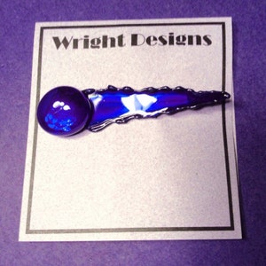 May include: A blue glass art piece with a rounded end and a jagged edge. The piece is on a white card with the text "Wright Designs" printed in black.