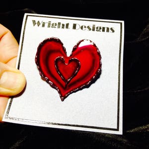 May include: A red heart-shaped pin with a smaller heart inside, held on a white card with the text "Wright Designs". The pin has a glossy finish and a dark outline. The overall design is a heart-themed accessory.