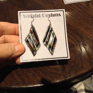 May include: A pair of silver dangle earrings with a diamond-shaped design. The earrings feature a dark brown and iridescent glass insert with a striped pattern. The earrings are displayed on a white card with the text "Wright Designs".