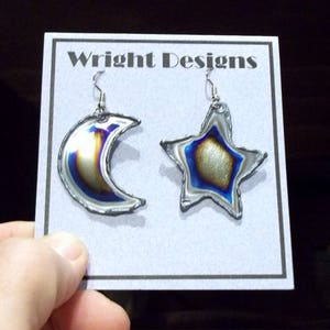 May include: A pair of handmade earrings featuring a crescent moon and a star. The earrings have a silver-tone metal outline with iridescent blue, gold, and silver coloring. The card behind the earrings reads "Wright Designs".