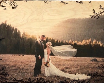 May include: A wedding photo of a couple standing in a field with a mountain range in the background. The bride is wearing a white dress and veil, and the groom is wearing a dark suit. The photo is printed on a wooden plaque with a burnt edge.