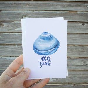 May include: A stack of white cards featuring a watercolor illustration of a blue seashell. The words "shell yeah!" are written in blue script below the shell. The cards are held against a weathered wood background.