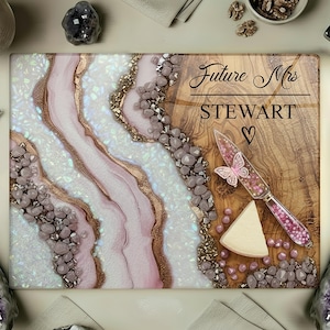 May include: A rectangular cutting board with a wood grain finish and a decorative resin design in pink, white, and gold. The text "Future Mrs STEWART" is displayed, along with a heart symbol. A cheese knife with a butterfly design is also present.