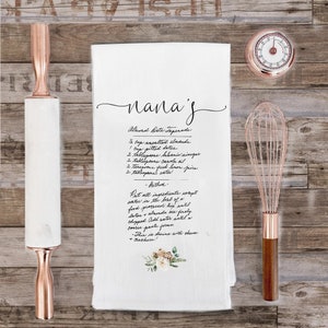 May include: A white kitchen towel with a handwritten recipe for Almond Date Tapenade. The recipe includes ingredients like almonds, dates, balsamic vinegar, canola oil, lemon juice, and water. The towel has a floral design in the bottom right corner.