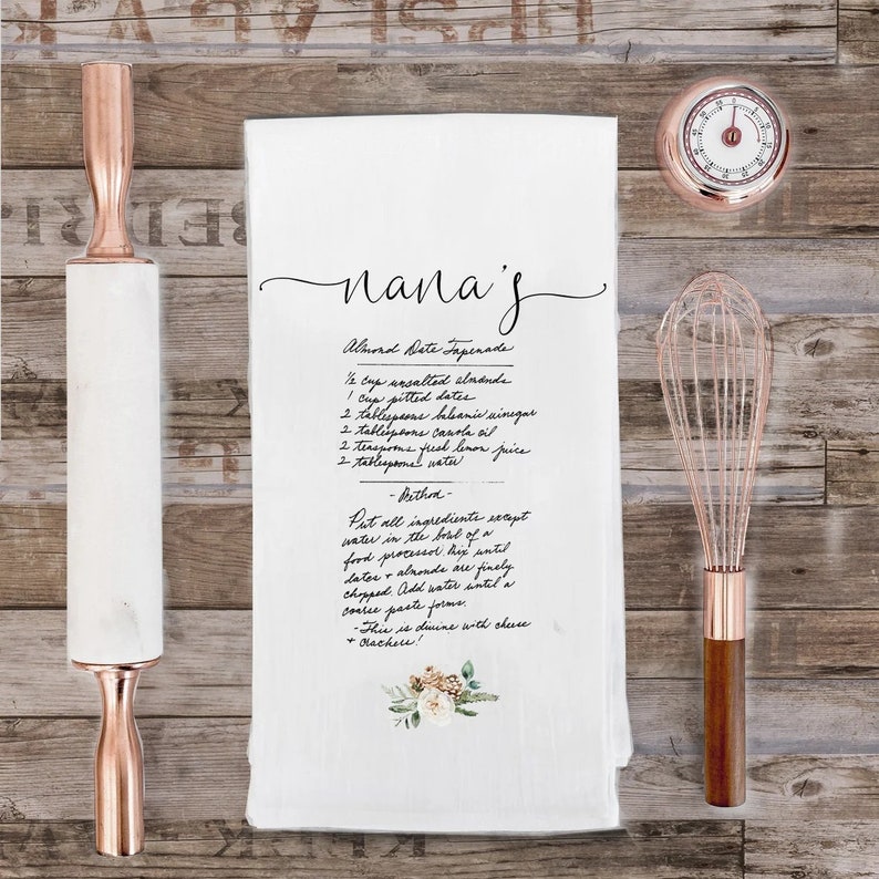 May include: A white kitchen towel with a handwritten recipe for Almond Date Tapenade. The recipe includes ingredients like almonds, dates, balsamic vinegar, canola oil, lemon juice, and water. The towel has a floral design in the bottom right corner.