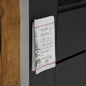 May include: A white kitchen towel with a red and white striped border. The towel features a watercolor illustration of a Christmas tree and presents. The text "Mom's" is written in cursive at the top of the towel, followed by "Mother's Eggnog (Charleston Style Recipe)". The recipe is written in cursive below the title.