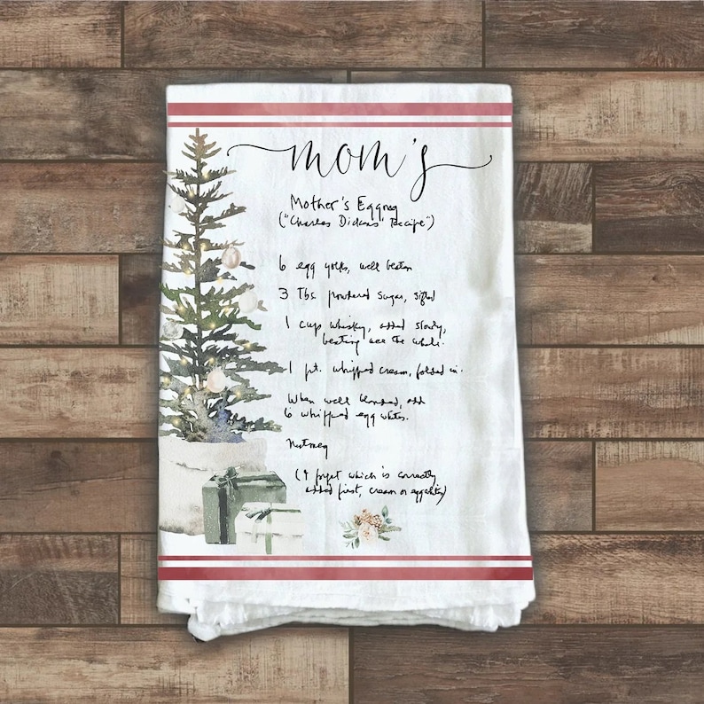 May include: A white kitchen towel with red stripes features a watercolor illustration of a Christmas tree with presents and a handwritten recipe for "Mother's Eggnog" (Charles Dickens' Recipe). The recipe includes ingredients like egg yolks, powdered sugar, whiskey, whipped cream, and nutmeg.