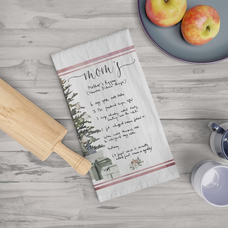 May include: A white tea towel with a watercolor illustration of a pine tree and a handwritten recipe for "Mother's Eggs" with the text "mom's" in pink script at the top.
