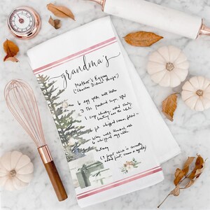 May include: A white kitchen towel with a pink and red stripe design. The towel features a watercolor illustration of a Christmas tree with presents and a handwritten recipe for "Mother's Eggnog" with the text "(Charles Didan's Recipe)".
