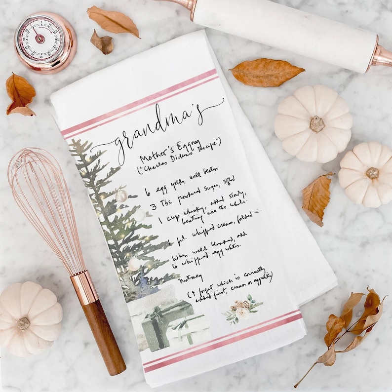 May include: A white kitchen towel with a pink and red stripe design. The towel features a watercolor illustration of a Christmas tree with presents and a handwritten recipe for "Mother's Eggnog" with the text "(Charles Didan's Recipe)".