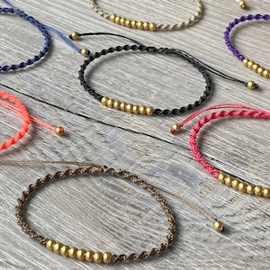 May include: A set of eight bracelets with gold beads and different coloured cords. The cords are in shades of pink, red, purple, blue, black, and brown.