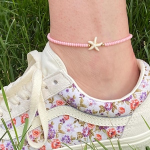 May include: Pink beaded anklet with a white starfish charm. The anklet is shown on a person's ankle with a floral patterned shoe.