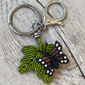 May include: A macramé keyring featuring a green leaf and a black butterfly with white and brown accents. The butterfly is attached to a green leaf, and the keyring has a silver ring and clasp.