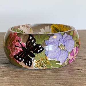 May include: A clear resin bangle bracelet with embedded dried flowers and a black and white butterfly. The bracelet features pink, purple, yellow, and white flowers. The bangle is on a wooden surface.