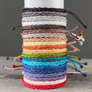 May include: A stack of 18 colourful braided bracelets in various colours, including black, white, brown, beige, orange, green, blue, purple, pink, and red. The bracelets are made of woven string and have a simple knot closure.