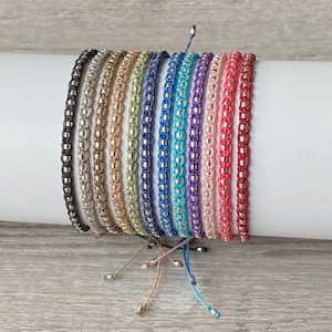 May include: A collection of 14 colourful braided bracelets with silver beads. The bracelets are arranged in a row on a white surface.