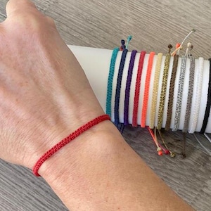 May include: A close-up of a red string bracelet on a person's wrist. A variety of colourful string bracelets are displayed in the background.