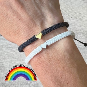 Peut inclure: Deux bracelets d'amitié avec des breloques en forme de cœur. Un bracelet est noir et l'autre est blanc. Les bracelets sont au poignet d'une personne. "Plus de couleurs disponibles" est écrit dans un arc-en-ciel en dessous des bracelets.