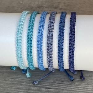 Peut inclure: Ensemble de six bracelets tressés dans des tons de bleu, allant du bleu clair au bleu foncé. Les bracelets sont faits d'un matériau doux et tissé et sont noués à l'extrémité.
