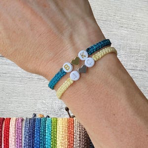 May include: A close-up of a wrist wearing two friendship bracelets. The bracelets are made of colourful string and have small white beads with letters and silver heart charms.