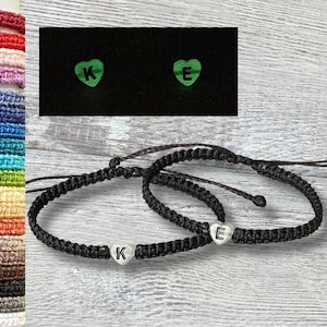 May include: Two black friendship bracelets with silver heart charms. The charms have the letters "K" and "E" on them. The bracelets are on a wooden surface.