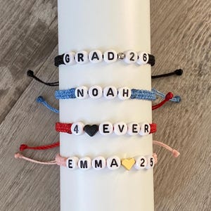 May include: Four handmade beaded bracelets with white letter beads spelling out "GRAD 26", "NOAH", "4 ♥ EVER", and "EMMA 25". Each bracelet has a different colour cord: black, blue, red, and pink. The bracelets are displayed on a white cylindrical object.