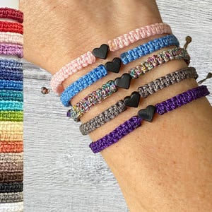 May include: A set of colourful friendship bracelets with black heart charms. The bracelets are made of woven string and are layered on a person's wrist.