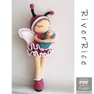 Op de afbeelding: Een handgemaakte gehaakte pop in een feeënontwerp, met een babypop. De pop draagt een bordeauxrode jurk met witte rand, een bijpassende hoed met antennes en roze schoenen. De baby is gewikkeld in een blauwgroene deken. De tekst "River Rice" is zichtbaar.