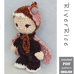 Könnte beinhalten: Eine Häkelpuppe mit cremefarbenem Gesicht, dunkelbraunem Kapuzenoutfit mit weißem Saum und rosafarbenen blattförmigen Flügeln. Die Puppe hat ein kleines Eichel-Accessoire an der Kapuze. Der Text lautet RiverRice crochet PDF pattern ENGLISH.