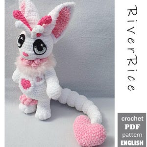 May include: A white and pink crocheted creature plush with large ears, a fluffy neck collar, a heart-shaped chest patch, and a long tail ending in a pink heart. The text RiverRice and crochet PDF pattern ENGLISH are visible on the right side.