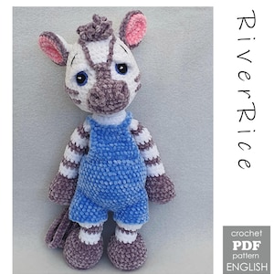 Op de afbeelding: Een gehaakte zebra knuffel met witte en grijze strepen, gekleed in een blauwe tuinbroek. De zebra heeft grote blauwe ogen, roze binnenoren en een grijze haarlok. De tekst RiverRice en crochet PDF pattern ENGLISH is aan de rechterkant zichtbaar.
