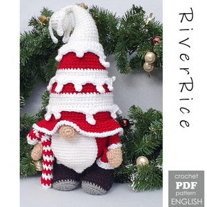 Op de afbeelding: Een handgemaakte gehaakte kabouterpop met een witte baard, rode jas en witte hoed. De kabouter houdt een zuurstok vast en heeft grijze en bruine schoenen. De afbeelding bevat de tekst "River Rice" en "crochet PDF pattern ENGLISH".