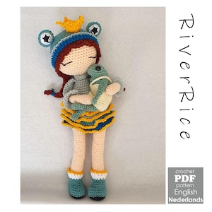 Op de afbeelding: Handgemaakte gehaakte pop met rood haar, een blauwe kikkerhoed met een gouden kroon, een grijze top en een gele en blauwe rok. De pop houdt een groene en crèmekleurige kikker vast. De tekst "River Rice" staat aan de rechterkant van de afbeelding.