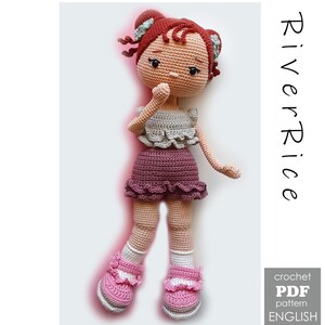 May include: A handmade crochet doll with red hair styled in two buns, wearing a gray top, a purple skirt, and pink shoes. The doll has a neutral expression. The image includes the text "River Rice" and "crochet PDF pattern ENGLISH".