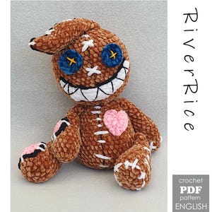 May include: A brown crocheted plush doll with a large head, one floppy ear, and blue button eyes with yellow stitching. It features a wide white stitched mouth, a pink heart patch on the chest, and white cross-stitch details. Text reads RiverRice crochet PDF pattern ENGLISH.