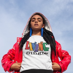 May include: A person wearing a white t-shirt with the text "LIVING single" in rainbow colors. The person is wearing a red jacket with the hood up and has black braids.