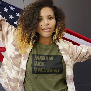 May include: A woman wearing a green t-shirt with the text "MADAM VICE PRESIDENT Kamala Harris" in black lettering. She is holding an American flag in each hand.