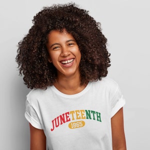 May include: A white t-shirt with a red, yellow, and green arc design that reads "Juneteenth 1865". The person wearing the shirt is smiling.