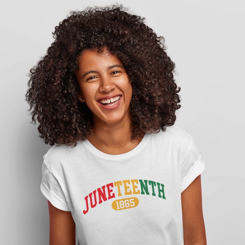 May include: A white t-shirt with a red, yellow, and green arc design that reads "Juneteenth 1865". The person wearing the shirt is smiling.