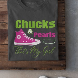 May include: A dark gray t-shirt with the text "chucks & pearls that's my girl" in green and pink. The shirt features a pink and white sneaker with a white star and a white pearl necklace.