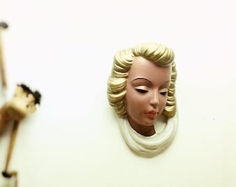 May include: A white wall with a wooden monkey figurine hanging from the ceiling and a white plaster face mask with blonde hair hanging on the wall.