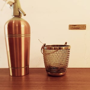 May include: A copper soda siphon with a green top and a brass wire mesh basket on a wooden surface.
