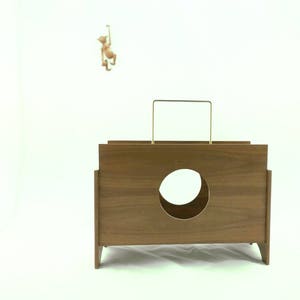 May include: A mid-century modern wooden box with a circular opening and a gold-colored handle. A small monkey figurine hangs from a thin rod above the box. The box is a warm brown color.